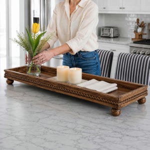 May include: A rectangular, wooden serving tray with a carved rope design around the edges and ball feet. Two lit, ivory-colored candles and a folded white cloth are on the tray. A clear vase with a yellow tulip and greenery is also on the tray.
