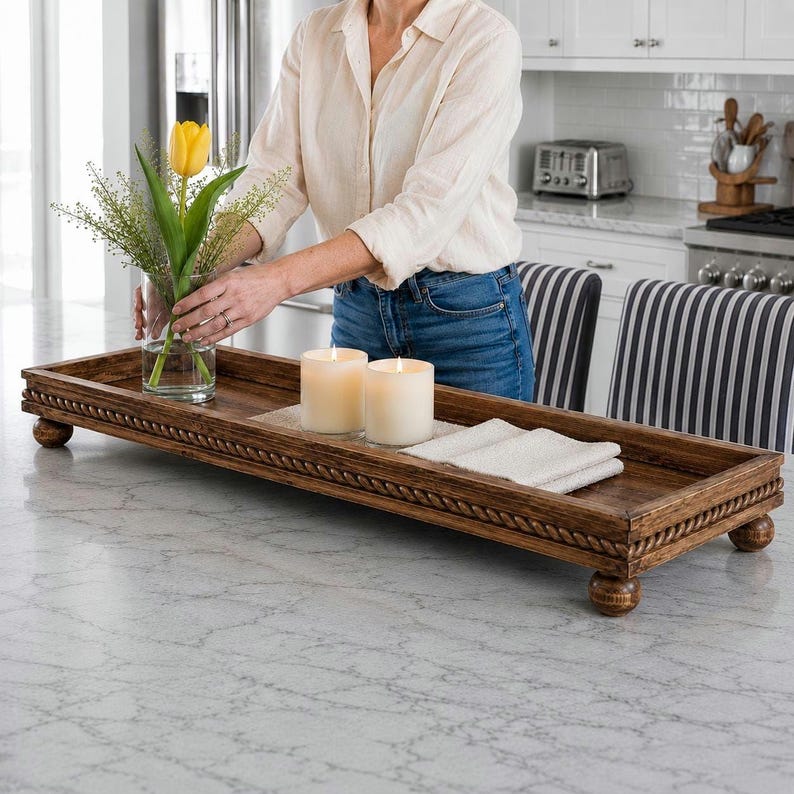 May include: A long, rectangular wooden tray with a rope-style carved border and four rounded feet. The tray holds a clear glass vase with a yellow tulip and green foliage, two white pillar candles, and a folded white cloth napkin on the surface.