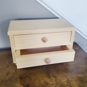 K&ouml;nnte beinhalten: Ein kleines Schubladenelement aus Holz mit zwei Schubladen. Die obere Schublade ist geschlossen und die untere Schublade ist offen. Die Schubladen haben Holzkn&ouml;pfe.