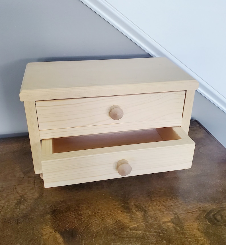 K&ouml;nnte beinhalten: Ein kleines Schubladenelement aus Holz mit zwei Schubladen. Die obere Schublade ist geschlossen und die untere Schublade ist offen. Die Schubladen haben Holzkn&ouml;pfe.