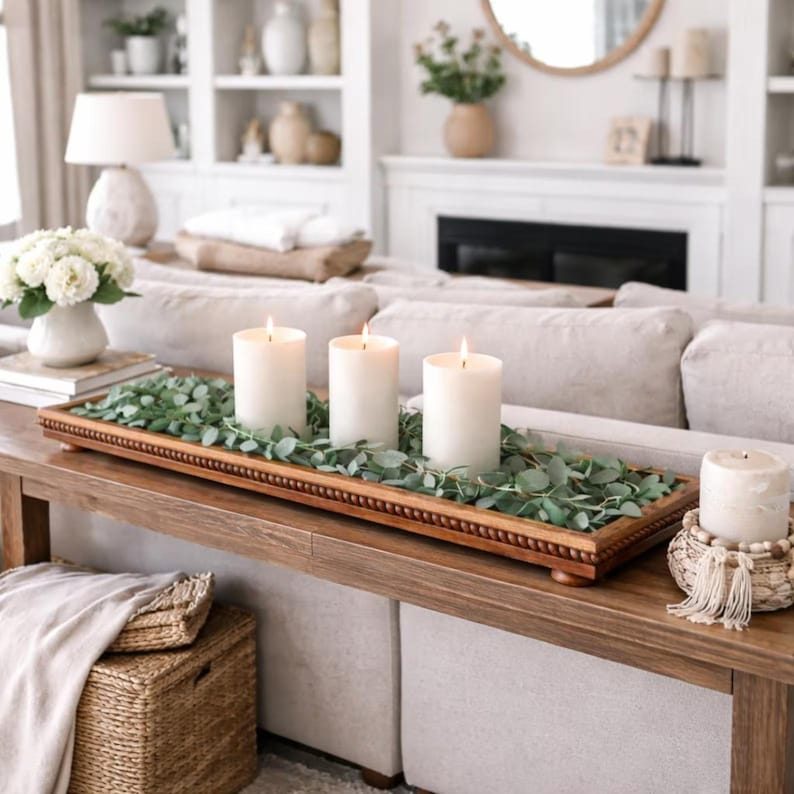 May include: A long rectangular wooden tray with a beaded edge and small feet, holding three white pillar candles nestled among green eucalyptus leaves. A separate small woven basket with a beaded rim and tassel trim holds a fourth pillar candle.