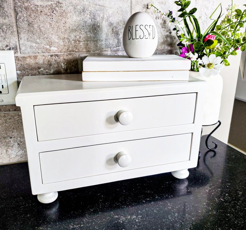 May include: A white wooden desktop organizer featuring two horizontal drawers with round knobs, supported by four small bun feet. Resting on top are two stacked white rectangular blocks and a white egg-shaped ornament with the word BLESSED.