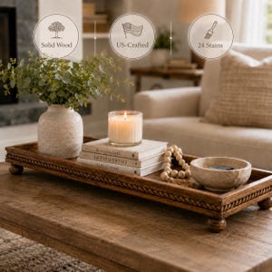 May include: A rectangular brown wooden tray with a rope-style border and four ball feet. The tray holds a textured beige vase with greenery, a lit candle in a glass jar, a stack of three books, a wooden bead garland, and a small stone bowl.