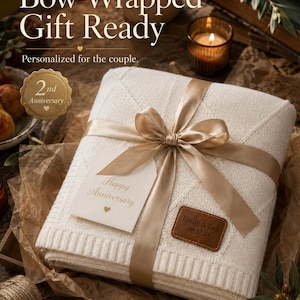 May include: A folded cream-colored knit blanket tied with a gold satin ribbon. A tag reads "Happy Anniversary" and a brown leather patch is embossed with "Emily & Jack 08.12.24". Text on the image includes "Bow-Wrapped Gift Ready" and "100% Cotton".