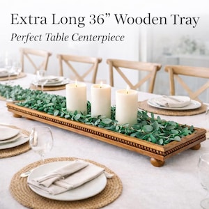 May include: A rectangular wooden tray measuring 36 inches in length, featuring a beaded edge and four small feet. The tray is filled with green eucalyptus leaves and three white pillar candles, positioned as a centerpiece on a dining table.