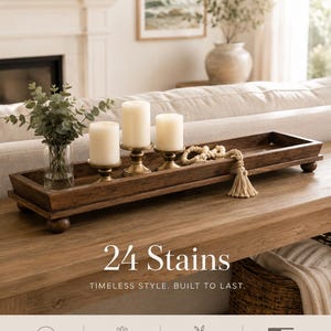 May include: A rectangular dark brown wooden tray with four ball feet, holding a clear glass vase with green foliage, three cream-colored pillar candles on gold-toned metal stands, and a cream-colored wooden bead garland with a tassel. Text reads 24 Stains.