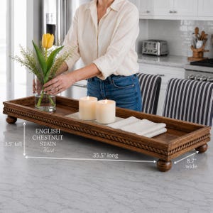 May include: A rectangular wooden tray with an English chestnut stain finish, featuring a rope-style border and four ball feet. The tray measures 35.5 inches long, 8.7 inches wide, and 3.5 inches tall, holding a glass vase with a yellow tulip.