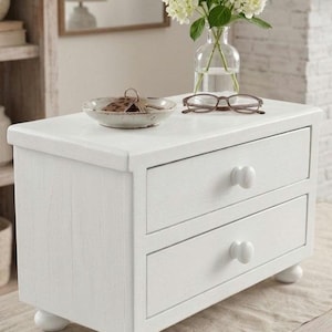 May include: A white wooden tabletop chest with two drawers, each featuring a round knob. The chest stands on four small bun feet. On top of the chest sits a small ceramic bowl containing keys, a pair of brown eyeglasses, and a clear glass vase.