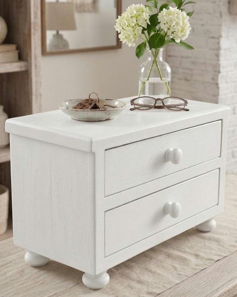 May include: A white wooden tabletop chest with two drawers, each featuring a round knob. The chest stands on four small bun feet. On top of the chest sits a small ceramic bowl containing keys, a pair of brown eyeglasses, and a clear glass vase.