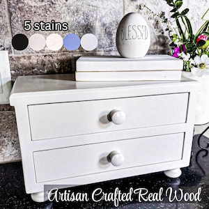 May include: A white wooden chest with two drawers, each featuring a round white knob. The chest stands on four small bun feet. Text on the image reads 5 stains and Artisan Crafted Real Wood. A white egg-shaped ornament with the word BLESSED is on top.
