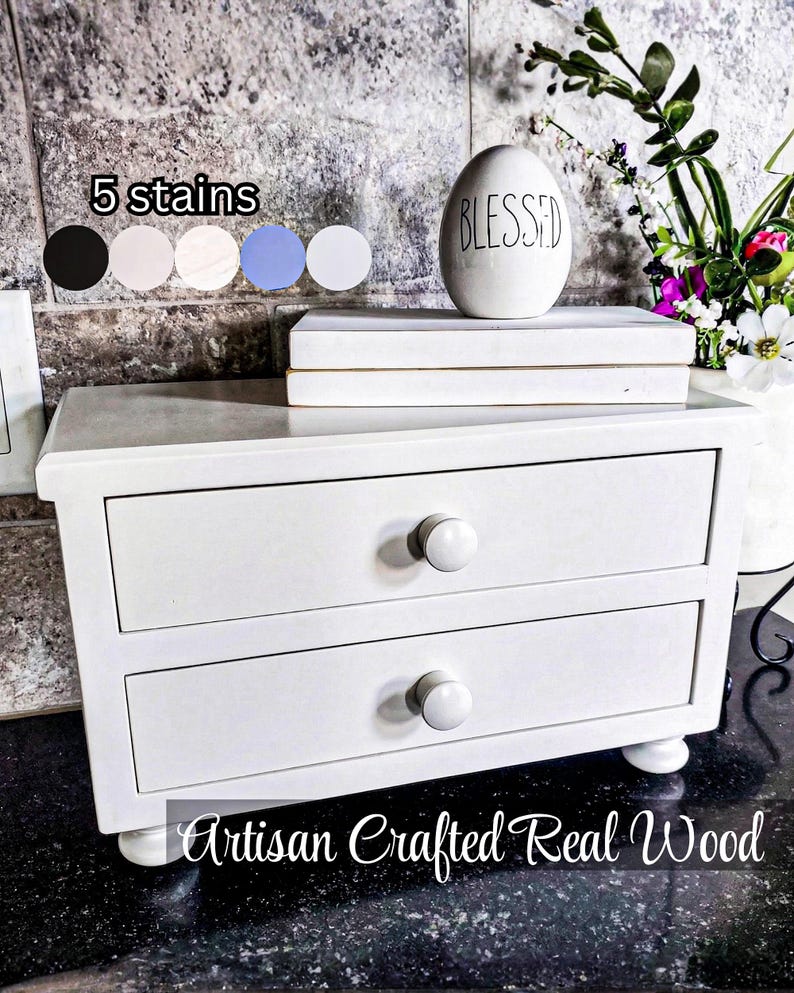 May include: A white wooden chest with two drawers, each featuring a round white knob. The chest stands on four small bun feet. Text on the image reads 5 stains and Artisan Crafted Real Wood. A white egg-shaped ornament with the word BLESSED is on top.