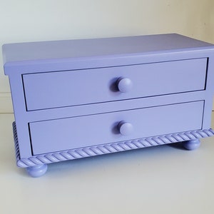May include: A rectangular lavender storage chest featuring two horizontal drawers with matching round knobs. The base of the chest is decorated with a carved rope-style trim and is supported by four small, rounded feet in the same solid color.