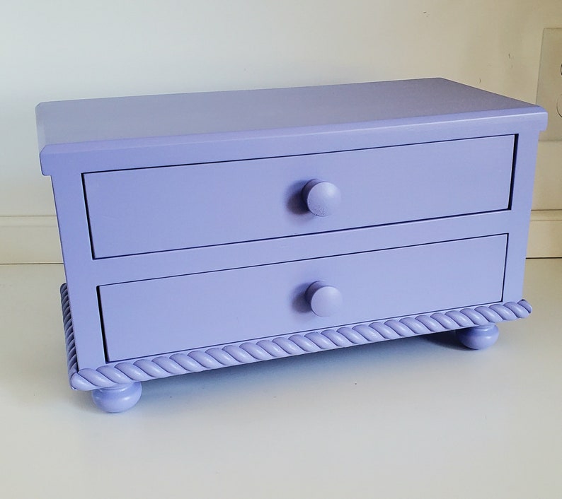 May include: A rectangular lavender storage chest featuring two horizontal drawers with matching round knobs. The base of the chest is decorated with a carved rope-style trim and is supported by four small, rounded feet in the same solid color.