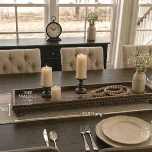 May include: A rectangular dark walnut stained wooden tray with a rope-style border and small feet. The tray measures 35.5 inches long, 8.75 inches wide, and 3.5 inches tall. It holds three pillar candles in black holders and a wooden bead garland.