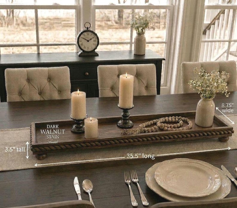 May include: A rectangular dark walnut stained wooden tray with a rope-style border and small feet. The tray measures 35.5 inches long, 8.75 inches wide, and 3.5 inches tall. It holds three pillar candles in black holders and a wooden bead garland.