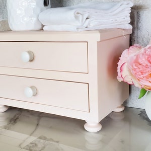May include: A light pink wooden desktop organizer featuring two horizontal drawers with simple white round knobs. The rectangular unit is supported by four small, rounded feet and has a smooth, matte finish across its entire exterior surface.