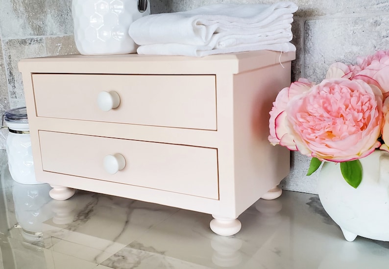 May include: A light pink wooden desktop organizer featuring two horizontal drawers with simple white round knobs. The rectangular unit is supported by four small, rounded feet and has a smooth, matte finish across its entire exterior surface.