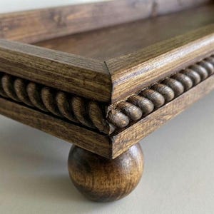 May include: A rectangular wooden tray with a dark brown stained finish. The outer edge of the tray features a decorative carved rope pattern. The tray is supported by four rounded ball-shaped feet, also finished in a matching dark brown wood stain.