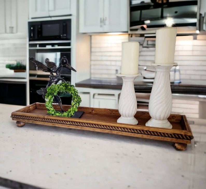 May include: A rectangular dark brown wooden tray with a rope-style border and small ball feet. The tray holds two white pillar candles in matching spiral-patterned candle holders and a small black metal bird figurine with a green wreath.