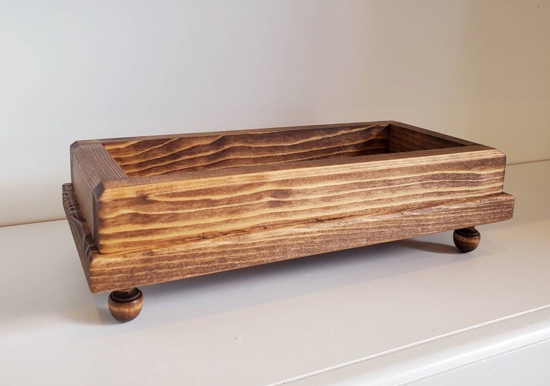 Pode incluir: Uma bandeja retangular de madeira, tingida de castanho escuro. Assenta em quatro pequenos p&eacute;s arredondados. O veio da madeira &eacute; vis&iacute;vel, adicionando textura. O design &eacute; simples e r&uacute;stico.
