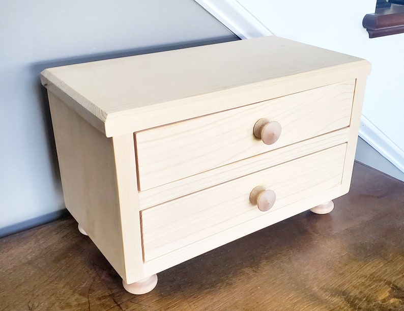 K&ouml;nnte beinhalten: Eine kleine Holzkiste mit zwei Schubladen und abgerundeten Holzkn&ouml;pfen. Die Kiste ist in einem hellen Beige gestrichen und hat eine nat&uuml;rliche Holzmaserung.