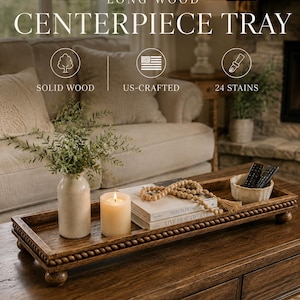 May include: A rectangular brown wooden tray with a beaded edge and four small feet. The tray holds a white ceramic vase with greenery, a lit pillar candle, two stacked books titled This Is Home and Live Beautifully, wooden beads, and a small bowl.