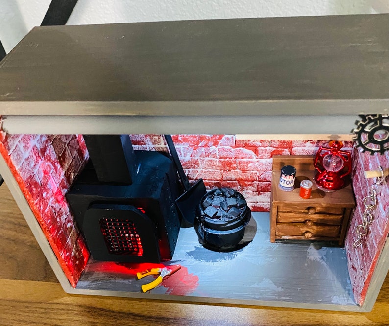 May include: A miniature diorama of a brick basement with a black wood-burning stove, a black metal bucket with coal, a small wooden dresser with a red can labeled "Motor", a red lantern, and a metal gear.
