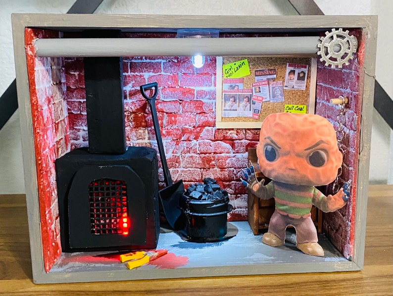 May include: A miniature diorama of a brick room with a black fireplace, a shovel, a bucket of coal, and a Freddy Krueger Funko Pop figure. The diorama has a light inside and a sign that says "Girl's Got a L.A.W.N." and "Call Cops!"