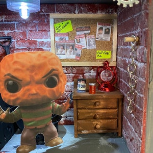 May include: A miniature diorama of a brick building with a red brick wall, a wooden door, and a wooden shelf with drawers. There is a small red lantern, a can of cola, and a can of motor oil on the shelf. A small figurine of Freddy Krueger, the character from the Nightmare on Elm Street movie series, stands in front of the building. The diorama is lit by a small light bulb. There are several missing person posters on the wall with the text "Girl 7401 Lakin", "Missing Person", "Missing", "Missing", "Wing Dog", "7890", and "Call Coal Co."