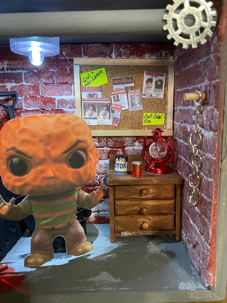 May include: A miniature diorama of a brick building with a red brick wall, a wooden door, and a wooden shelf with drawers. There is a small red lantern, a can of cola, and a can of motor oil on the shelf. A small figurine of Freddy Krueger, the character from the Nightmare on Elm Street movie series, stands in front of the building. The diorama is lit by a small light bulb. There are several missing person posters on the wall with the text "Girl 7401 Lakin", "Missing Person", "Missing", "Missing", "Wing Dog", "7890", and "Call Coal Co."