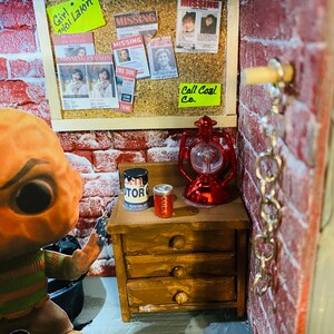 May include: A miniature dollhouse room with a red brick wall, a corkboard with missing person posters and a note that says "Girl 2401 Lakin", a wooden dresser with a can of cola and a can of paint, a red lantern, and a chain hanging from the wall.