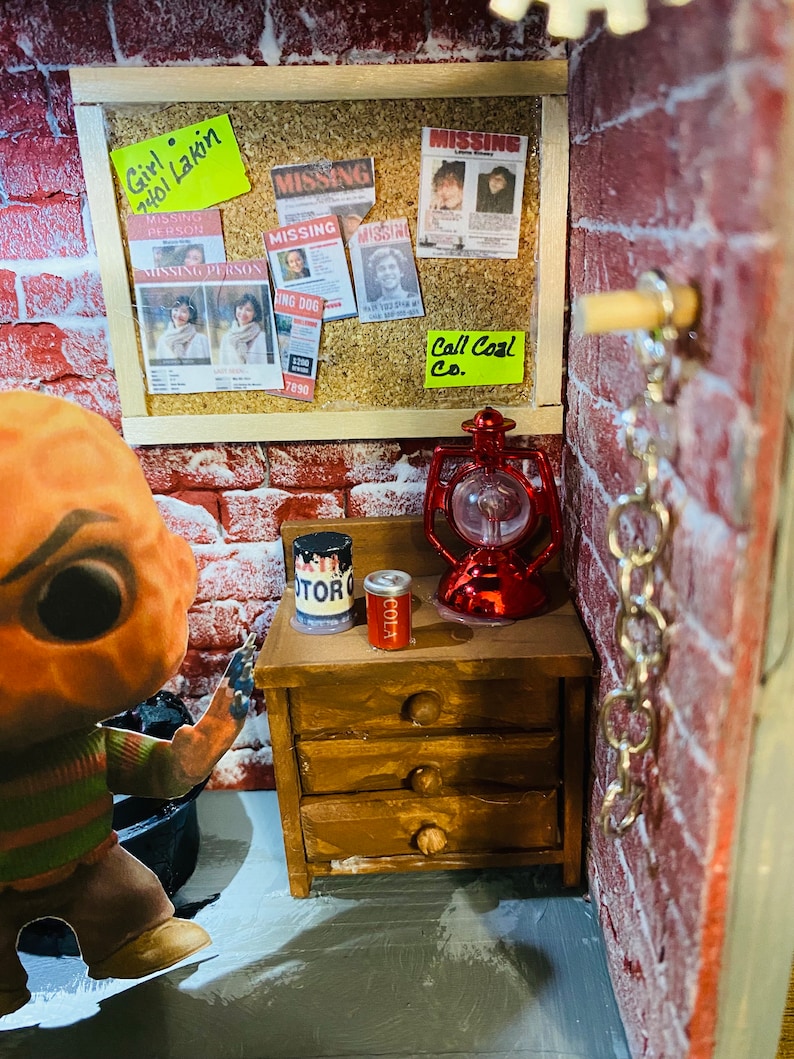May include: A miniature dollhouse room with a red brick wall, a corkboard with missing person posters and a note that says "Girl 2401 Lakin", a wooden dresser with a can of cola and a can of paint, a red lantern, and a chain hanging from the wall.