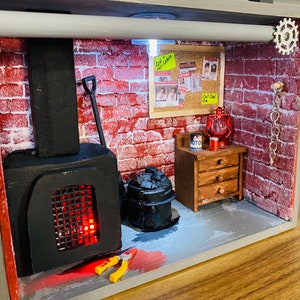 May include: A miniature diorama of a basement with a brick wall, a black wood-burning stove, a metal bucket with coal, a small wooden table with drawers, a red lantern, a shovel, a pair of pliers, and a yellow sticky note that says "Girl,  No Light!"
