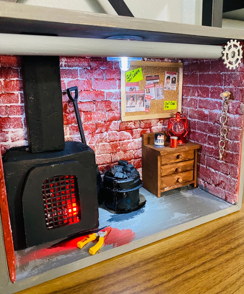 May include: A miniature diorama of a basement with a brick wall, a black wood-burning stove, a metal bucket with coal, a small wooden table with drawers, a red lantern, a shovel, a pair of pliers, and a yellow sticky note that says "Girl,  No Light!"