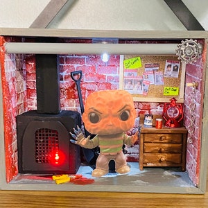 May include: A miniature diorama of a brick room with a fireplace, a small wooden dresser, and a Funko Pop figure of a character from the horror movie "The Thing". The diorama is lit from within, and the figure is wearing a striped shirt and a red mask.