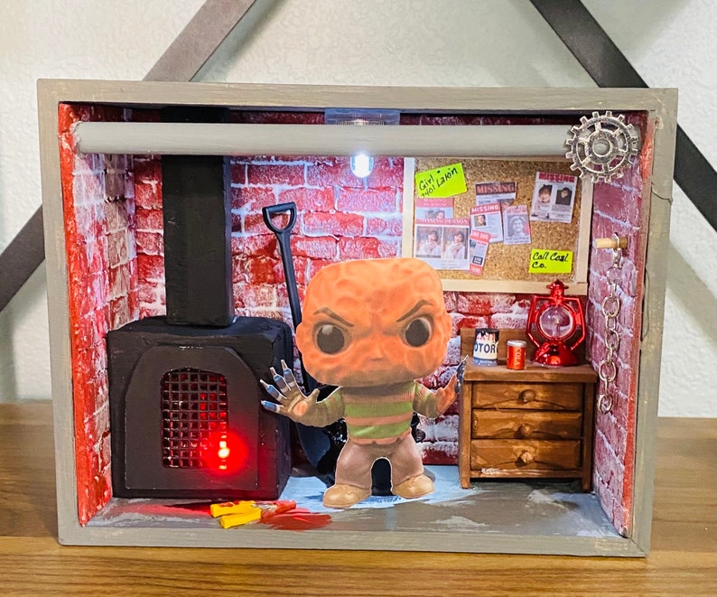 May include: A miniature diorama of a brick room with a fireplace, a small wooden dresser, and a Funko Pop figure of a character from the horror movie "The Thing". The diorama is lit from within, and the figure is wearing a striped shirt and a red mask.