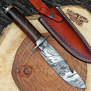 Peut inclure: Un couteau de chasse en acier de Damas avec un manche en bois foncé et un étui en cuir. La lame présente un motif unique et l'étui est marron avec des coutures blanches. Le couteau est posé sur une surface en bois.