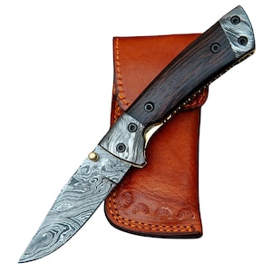 Peut inclure: Un couteau pliant avec une lame en acier Damas au motif ondulé gris et argent, un manche en bois foncé avec des mitres en métal et un bouton de pouce en laiton. Le couteau est posé sur un étui en cuir beige avec des coutures visibles.