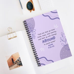 May include: A spiral-bound notebook with a light purple cover featuring dark purple abstract wavy lines, leaf illustrations, and the text: And see that ye have faith, hope, and charity, and then ye will always ABOUND in good works Alma 7:23-24.