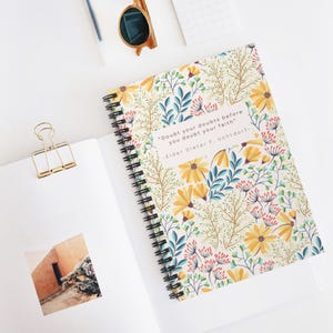 May include: A spiral-bound notebook featuring a floral pattern of yellow flowers and blue leaves with the text "Doubt your doubts before you doubt your faith" by Elder Dieter F. Uchtdorf. A gold binder clip and sunglasses are visible nearby.