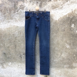 Op de afbeelding: Een donkerblauwe spijkerbroek hangt aan een houten hanger tegen een getextureerde grijze muur.