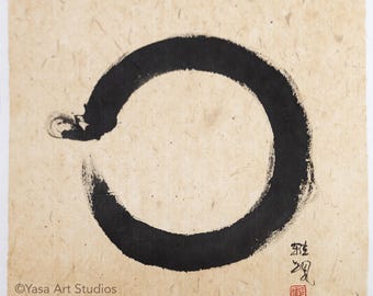 May include: A square piece of textured, light tan paper featuring a hand-painted black ink circle, known as an enso. The artwork includes black calligraphy characters and a small red square seal in the bottom right corner, with text at the bottom.