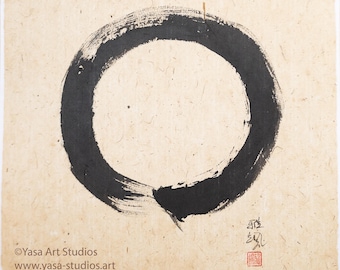 May include: A black ink ensō circle painted on textured, off-white paper. The brushstroke forms a complete circle with a slight opening. Japanese characters and a red seal are in the lower right corner. The text "Yasa Art Studios" and a website address are in the lower left corner.