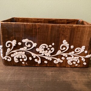 May include: A rectangular wooden planter with a white floral design. The planter has a dark brown stain and a white glaze.
