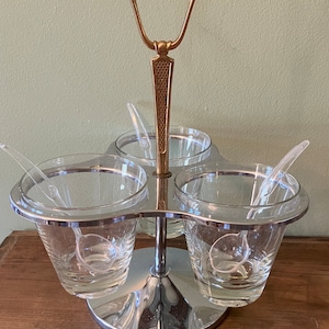 May include: A chrome and gold condiment server with three glass bowls and spoons. The server has a handle on top.