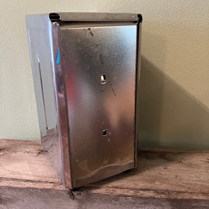 May include: A silver metal napkin holder with a slightly curved front edge. The holder has a rectangular shape and is made of a shiny metal.