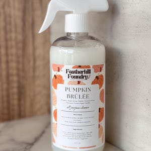 May include: A clear plastic spray bottle containing a milky white liquid, topped with a white trigger sprayer. The label features orange pumpkin illustrations and the text Featherhill Foundry, PUMPKIN BRÛLÉE, all purpose cleaner, 16 fl oz.