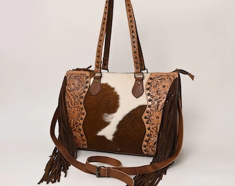 May include: A brown and white cowhide tote bag with a long, adjustable shoulder strap. The bag features decorative floral tooling on the sides, fringe, and studded accents on the handles. The bag measures approximately 14 inches wide.
