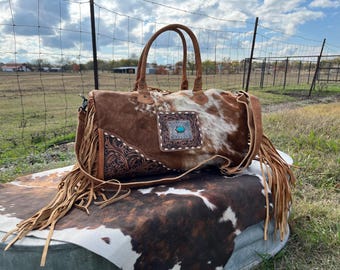 May include: A brown and white cowhide leather duffel bag with fringe detailing. The bag has a turquoise stone accent and a tooled leather design.