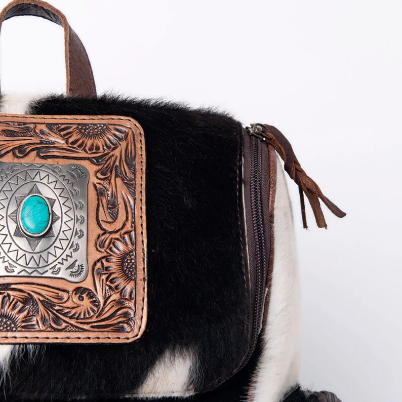 Op de afbeelding: Een zwart-witte koeienhuid rugzak met een bruine leren klep en een zilveren en turquoise concho. De rugzak heeft een ritssluiting en een bruine leren band.